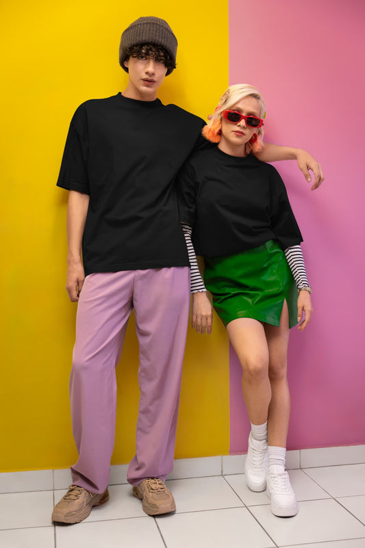 Two people standing against a yellow and pink wall, wearing black tops and colorful pants.