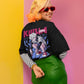 Person wearing a black jacket with a graphic design, striped shirt, and green skirt against a pink and yellow background.