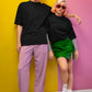 Two people standing against a yellow and pink wall, wearing black tops and colorful pants.